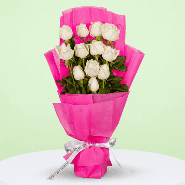 Bouquet of 12 white roses with green leaves, wrapped in bright fuchsia pink non-woven paper and tied with a white and black ribbon.