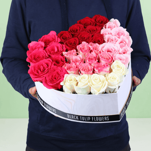 A vibrant combination of red, pink, and white roses arranged in a white heart-shaped box, topped with a golden "Love You" topper