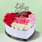 A vibrant combination of red, pink, and white roses arranged in a white heart-shaped box, topped with a golden "Love You" topper