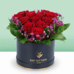 A black box featuring red roses in heart shape with purple filler flowers and green foliage.