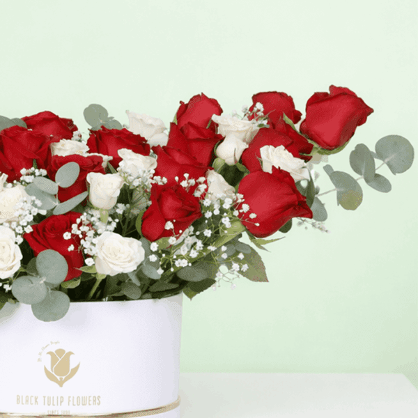 A white round flower box filled with red roses, white spray roses, baby’s breath, and eucalyptus.