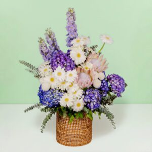 Grand flower basket arrangement with protea, hydrangea, delphinium, ping pong blooms, gerberas, and eucalyptus in a round basket.