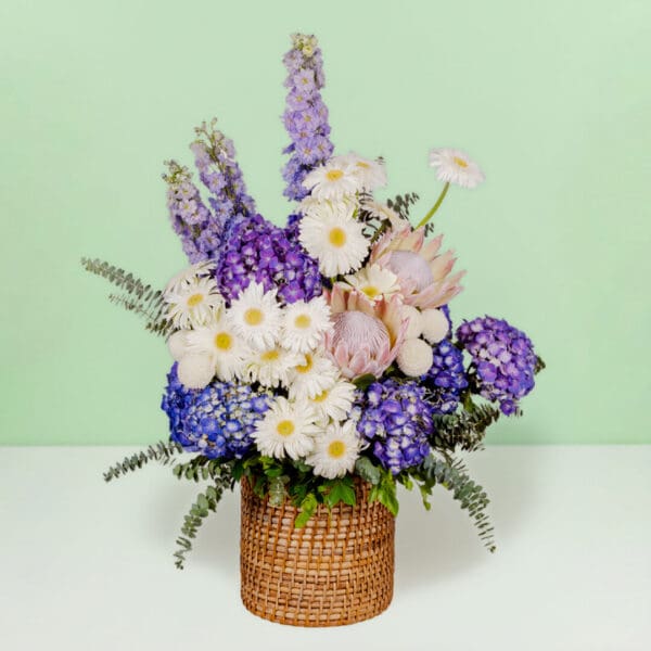 Grand flower basket arrangement with protea, hydrangea, delphinium, ping pong blooms, gerberas, and eucalyptus in a round basket.