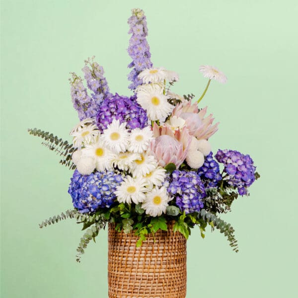 Grand flower basket arrangement with protea, hydrangea, delphinium, ping pong blooms, gerberas, and eucalyptus in a round basket.